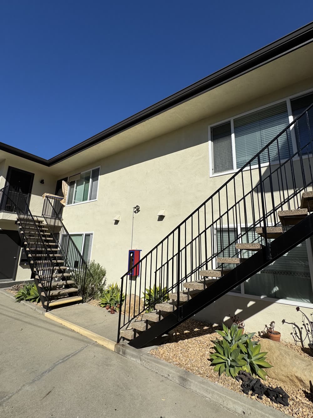 A two-story cream apartment building with black steel exterior stairs and railings under a blue sky.