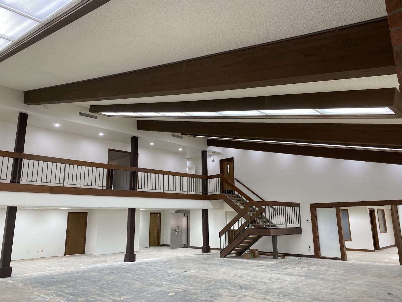 A large commercial interior with bright white walls, dark wood beams and posts, a mezzanine with a railing and an open wood staircase.
