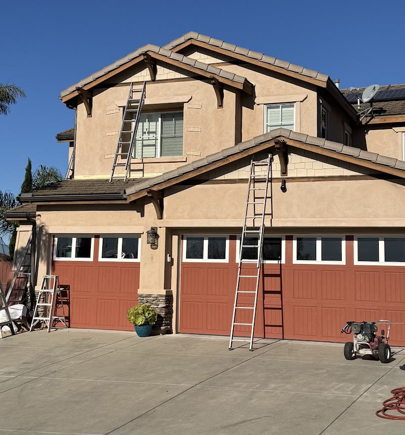 The same stucco house from the driveway, with extension ladders up on both gables and against the garage, step ladders and a pressure washer set out.