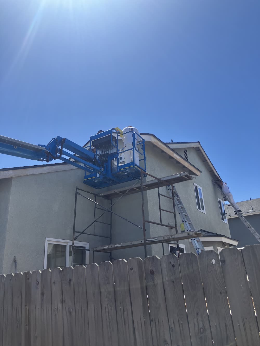 Two painters in white at work on a gray two-story stucco house, one in a blue boom lift over scaffolding and one on an extension ladder at the eave.