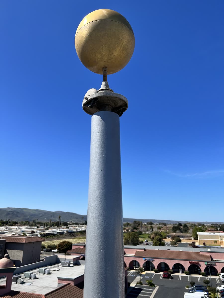 A gray-painted metal pole topped by a gold ball, seen from a rooftop high above a shopping center and the hills beyond.