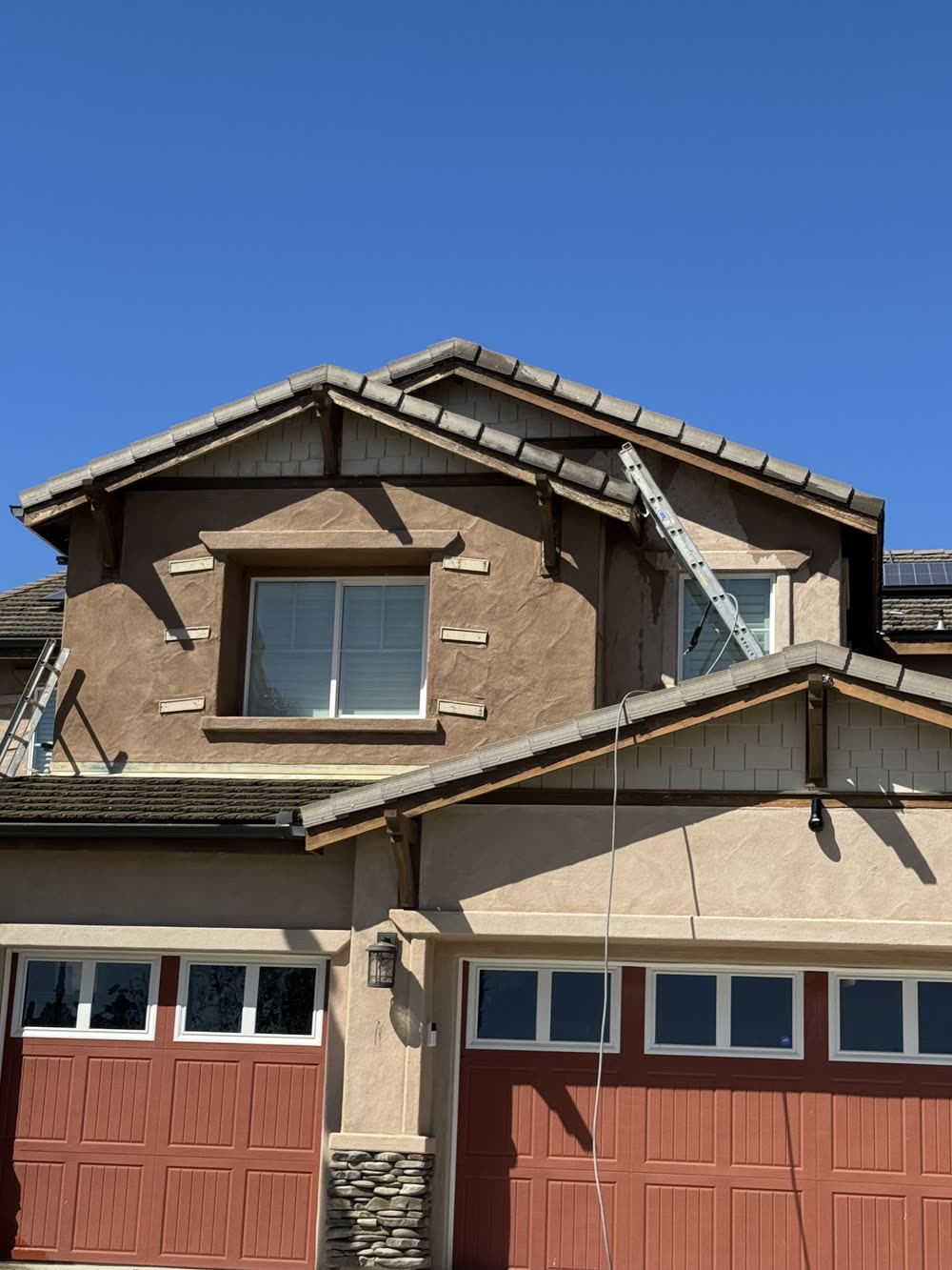 Close view of a stucco house's gables with the fascia and rafter tails stripped to bare wood, an extension ladder against the upper wall and red garage doors below.