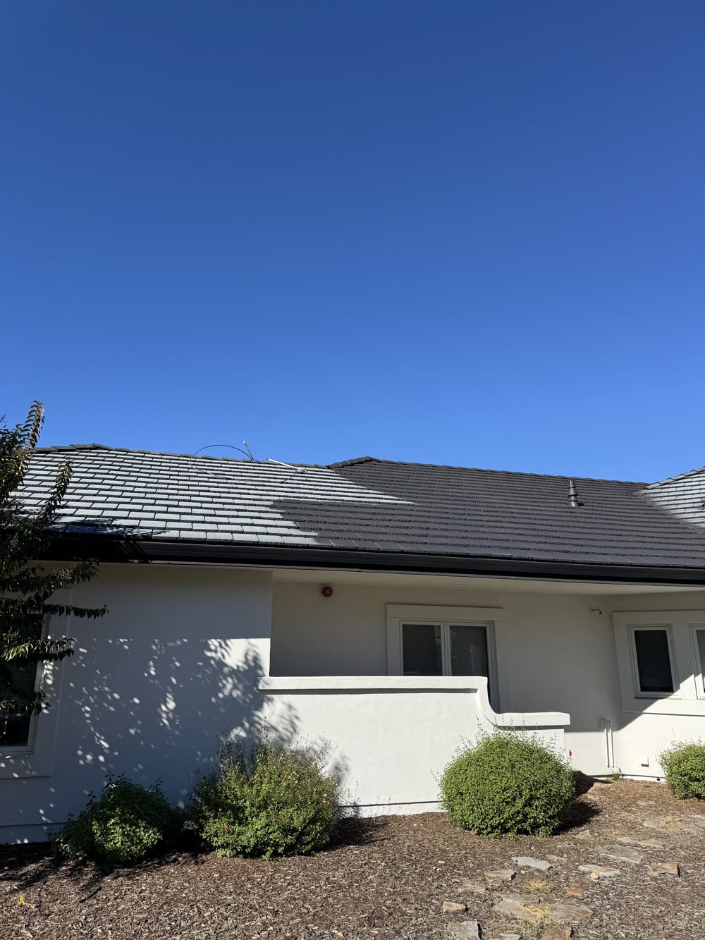 A single-story white stucco house with a dark tile roof and low shrubs in a bark bed.